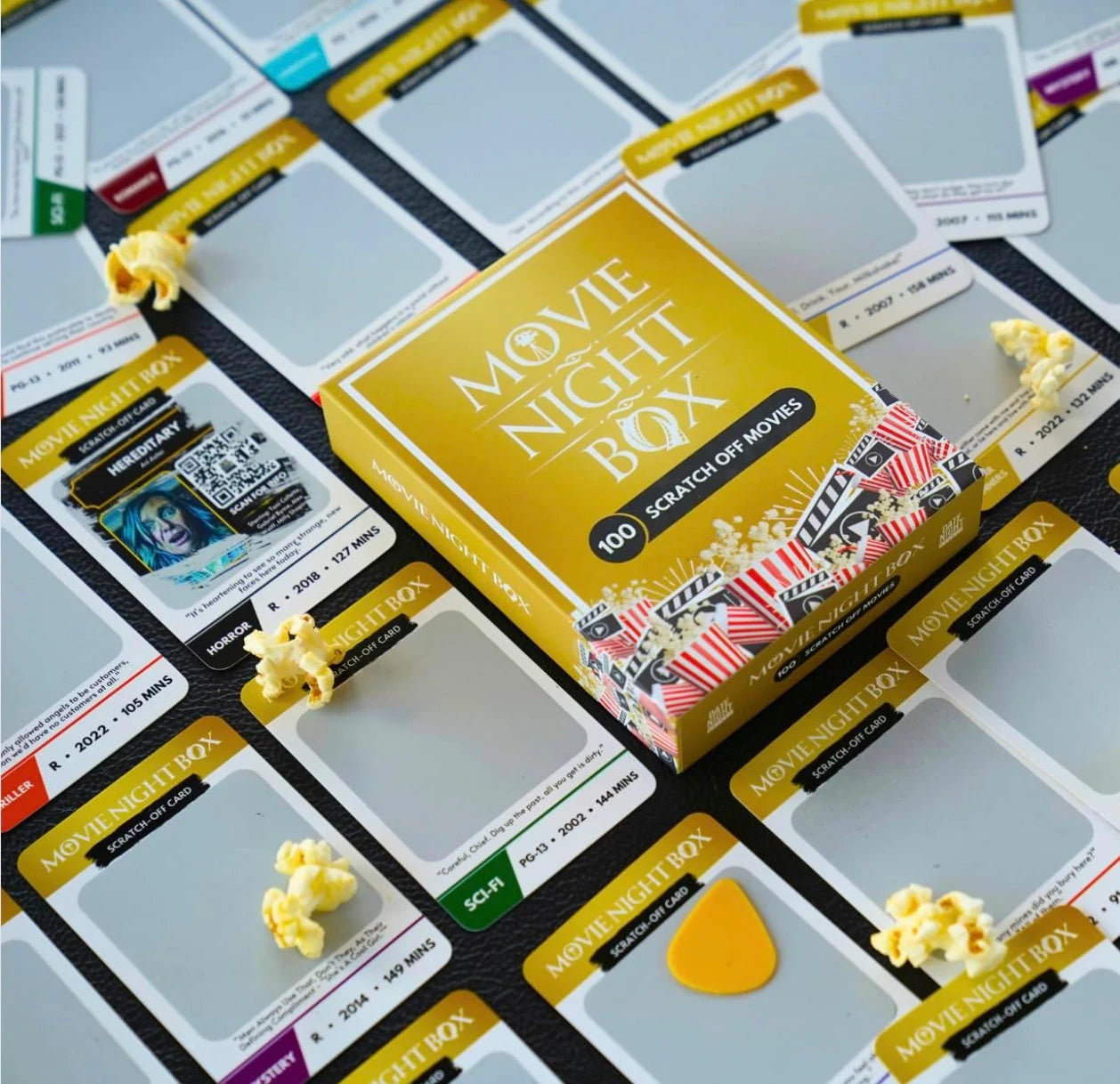 Movie Night Box from Date Night Brands featuring 100 scratch-off movie cards spread out on a table, showcasing genres like horror, sci-fi, and comedy, with a QR code for movie details, surrounded by popcorn for a cinematic experience.